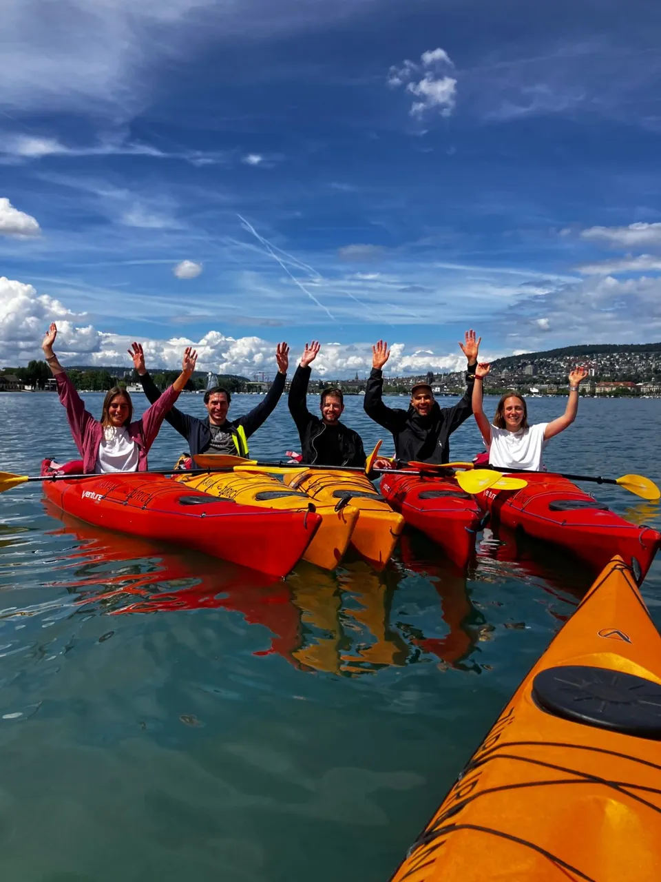 Fünf Personen aus dem Loopia-Team in roten und gelben Kajaks auf dem Zürichsee, die Arme in der Luft, dahinter die Stadt Zürich